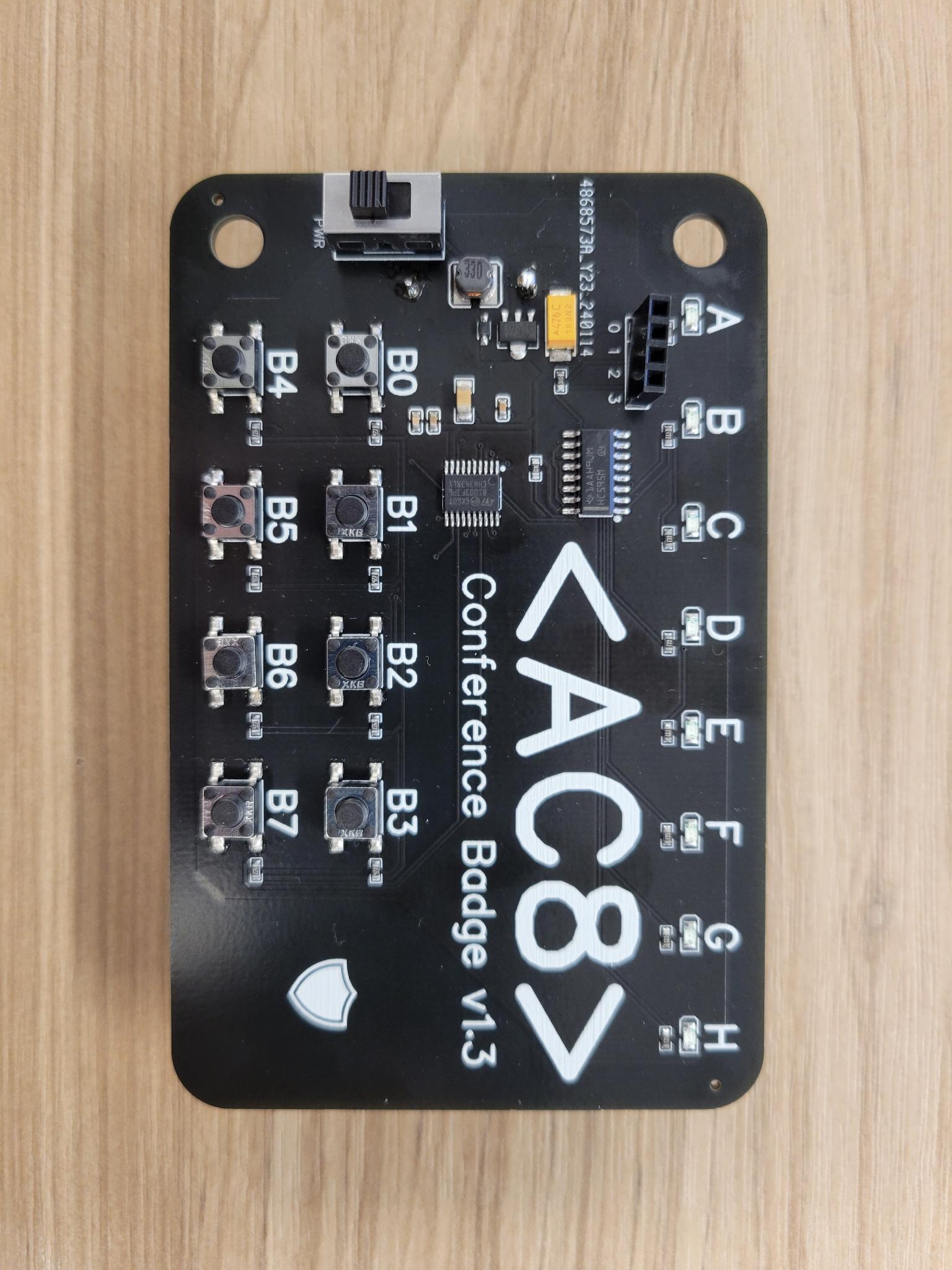 A close-up of the badge: A black PCB labelled AC8 / Conference Badge v1.3